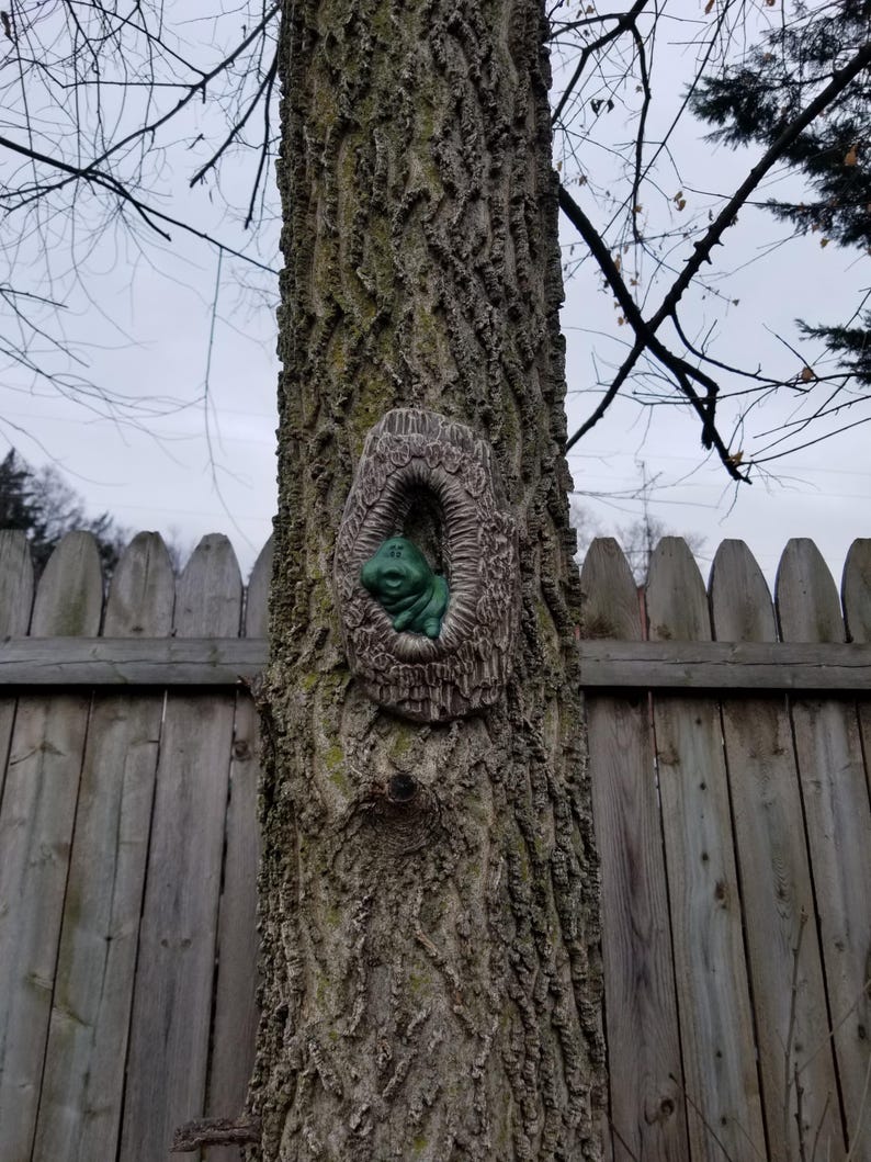 May include: A tree trunk with a decorative, oval-shaped sculpture of a frog in a hollow. The frog is green, and the sculpture has a textured, brown, bark-like appearance. A wooden fence and bare tree branches are in the background.