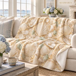 Puede incluir: Una manta de felpa con un estampado detallado de varias conchas marinas, dólares de arena y estrellas de mar en tonos crema, beige y dorado. La manta tiene una superficie suave y texturizada, colocada sobre un sofá con un diseño costero.