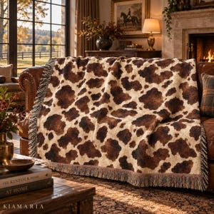 May include: A woven throw blanket featuring a brown and cream cow print pattern. The blanket has a textured surface and is finished with fringed edges along the sides. The design consists of irregular dark brown patches on a light cream background.