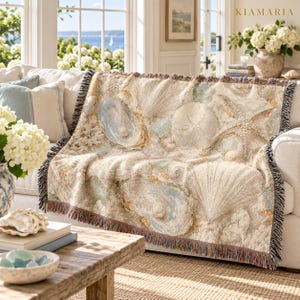 May include: A woven throw blanket featuring a detailed coastal pattern of seashells, starfish, and sand dollars in shades of beige, cream, and soft blue. The blanket has a fringed edge and the text KIAMARIA is visible in the upper right corner.