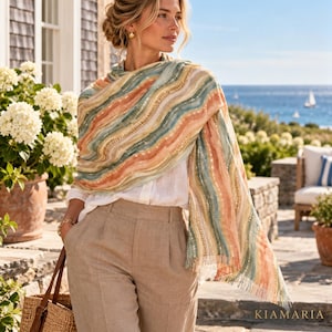 May include: A lightweight scarf featuring a wavy, multi-colored pattern in shades of peach, teal, and cream with subtle metallic gold accents. The scarf is draped over a white shirt and beige trousers, with the brand name KIAMARIA visible.