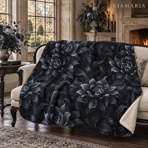 May include: A plush throw blanket featuring a dark, moody floral pattern of layered black and charcoal gray dahlia-like blossoms. The blanket has a soft, cream-colored sherpa fleece lining visible at the folded corner, draped over a sofa.
