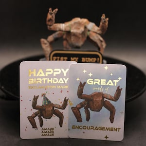 May include: Two rectangular greeting cards with gold foil text. The left card reads HAPPY BIRTHDAY EXCLAMATION MARK with a crab wearing a party hat. The right card reads GREAT words of ENCOURAGEMENT with a crab and gold star accents.