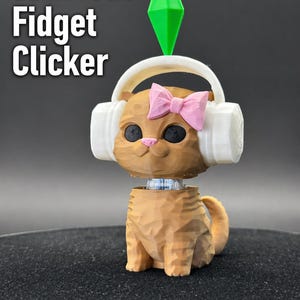 Puede incluir: Un fidget clicker de gato marrón impreso en 3D con auriculares blancos y un lazo rosa. El gato tiene ojos negros y un diamante verde encima de la cabeza. El texto "Sims Cat Fidget Clicker" se muestra en blanco.