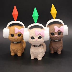 May include: Three cat figurines in shades of brown, tan, and dark brown, each wearing white headphones and a pink bow. Each cat has a colored diamond shape above its head. The figurines have a collar and are on a black surface.