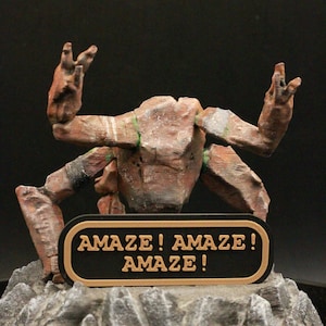 May include: A brown, stylized creature sculpture with outstretched arms and a sign that reads "AMAZE! AMAZE! AMAZE!". The sculpture is mounted on a rocky base, with a black background.