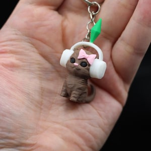 May include: A brown cat figurine keychain with white earmuffs and a pink bow. The cat has black eyes and a green diamond-shaped charm. The keychain is made of a metal ring and chain.