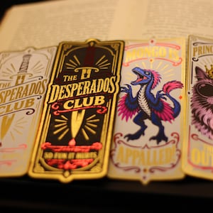Könnte beinhalten: Vier rechteckige Lesezeichen mit Goldfolienakzenten. Zwei zeigen ein Schwert und den Text The Desperados Club. Eines zeigt einen blauen und rosa Dinosaurier mit dem Text Mongo Is Appalled. Eines zeigt eine Katze mit Krone und Sonnenbrille.