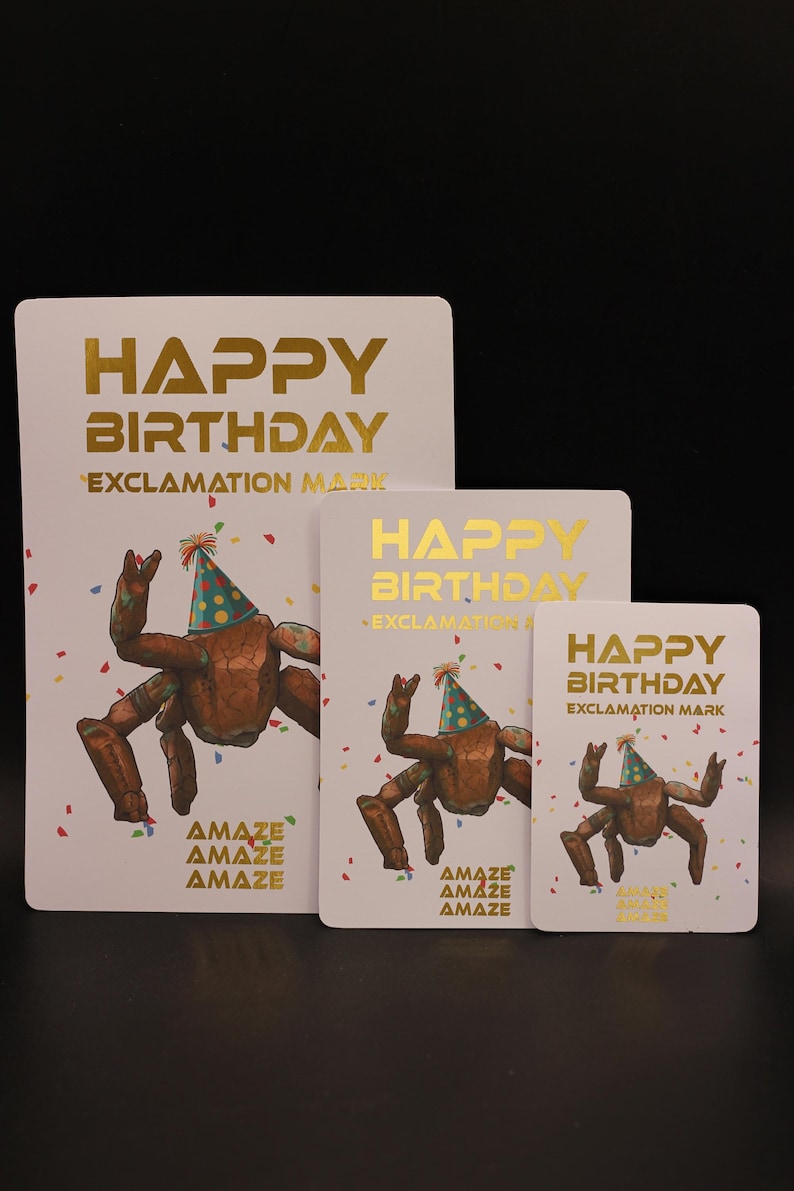May include: Three white rectangular cards of varying sizes featuring gold foil text that reads HAPPY BIRTHDAY EXCLAMATION MARK. Each card displays a brown crab wearing a colorful polka dot party hat surrounded by scattered confetti and the word AMAZE.