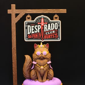 May include: A brown cat figurine wearing a gold crown, purple circular glasses, and a purple cape with a gold chain, sitting on a purple tufted cushion. Above the cat hangs a sign reading DESPERADO CLUB SO FUN IT HURTS with a red sword graphic.