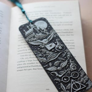 Peut inclure: Marque-page noir avec des images en relief d'un château, d'un hibou, d'une fiole de potion et d'autres éléments fantastiques. Une gland turquoise est attaché. Le marque-page est posé sur un livre ouvert.