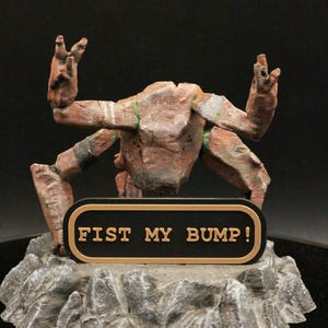 May include: A brown, multi-limbed creature figurine with a textured, rocky base. The figure features a black rectangular plaque with a gold border and the text FIST MY BUMP! in gold lettering. The creature has raised arms and a weathered appearance.
