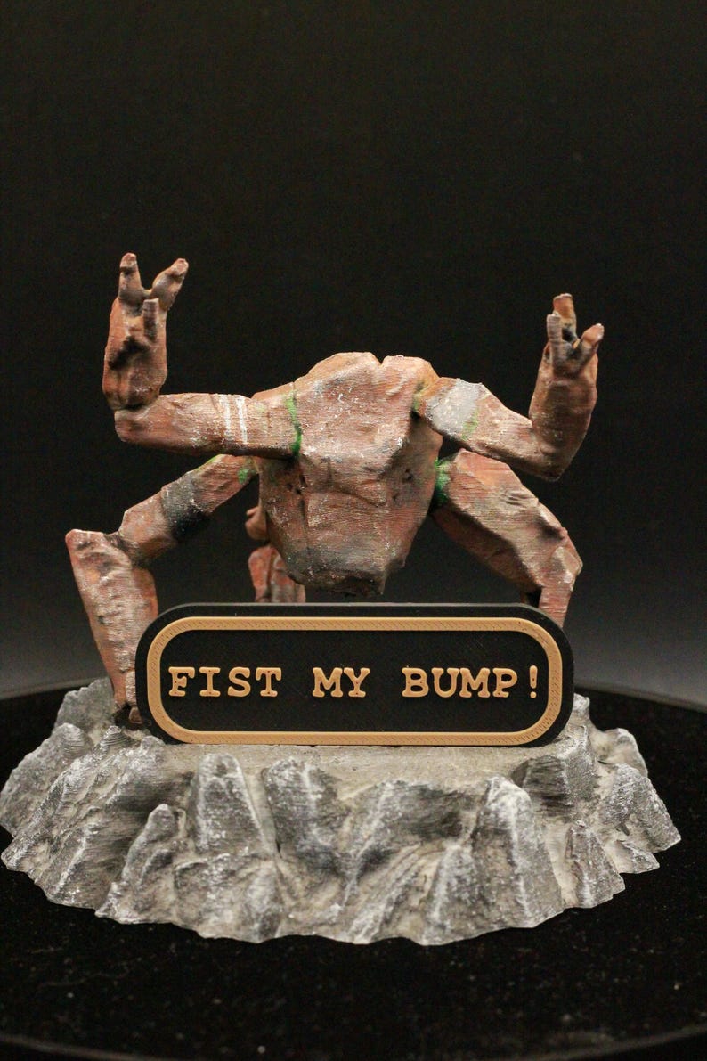 May include: A brown, multi-limbed creature figurine with a textured, rocky base. The figure features a black rectangular plaque with a gold border and the text FIST MY BUMP! in gold lettering. The creature has raised arms and a weathered appearance.