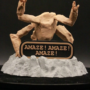 May include: A tan-colored, multi-limbed creature figurine with a textured surface, positioned behind a black, oval-shaped plaque with a tan border. The plaque features the text "AMAZE! AMAZE! AMAZE!" in tan lettering. The figure rests on a gray, rocky base.
