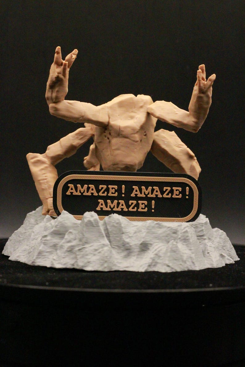 May include: A tan-colored, multi-limbed creature figurine with a textured surface, positioned behind a black, oval-shaped plaque with a tan border. The plaque features the text "AMAZE! AMAZE! AMAZE!" in tan lettering. The figure rests on a gray, rocky base.