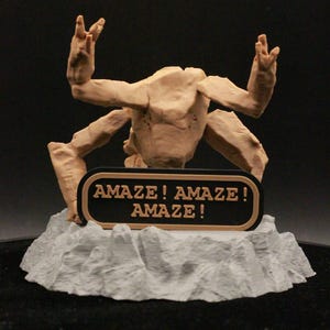 May include: A tan-colored, multi-limbed creature figurine with a textured surface, positioned on a jagged, light gray rocky base. A black rectangular sign with a tan border is attached to the front, featuring the text AMAZE! AMAZE! AMAZE!