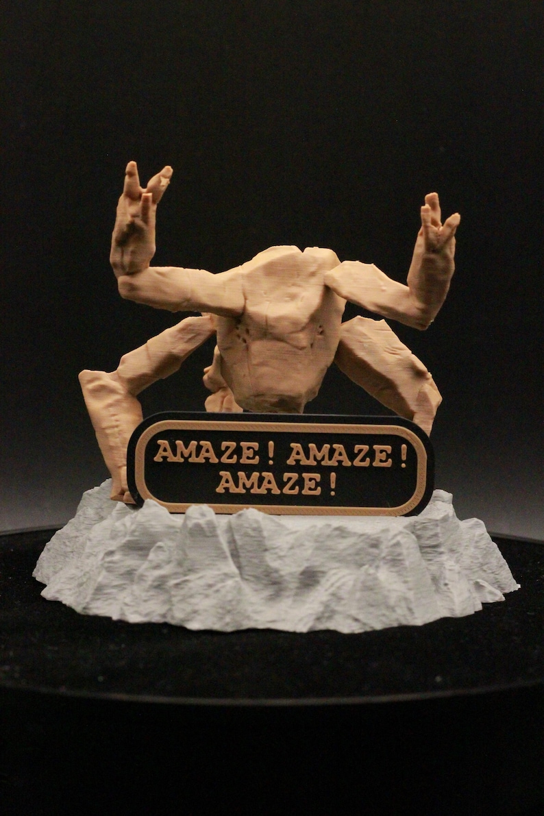 May include: A tan-colored, multi-limbed creature figurine with a textured surface, positioned on a jagged, light gray rocky base. A black rectangular sign with a tan border is attached to the front, featuring the text AMAZE! AMAZE! AMAZE!