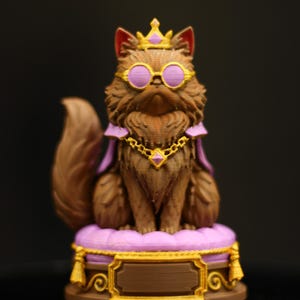 Könnte beinhalten: Eine braune Katzenfigur mit goldener Krone, rundem Brillenmodell mit violettem Rand und einem violetten Umhang mit goldener Kette. Die Katze sitzt auf einem violetten, gesteppten Kissen auf einem braun-goldenen Sockel mit Namensschild.