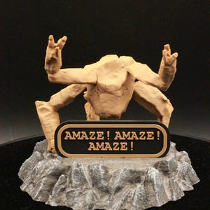 May include: A tan-colored, multi-limbed creature figurine with a textured, rocky base. A black, oval-shaped plaque is attached to the front of the base, featuring the text "AMAZE! AMAZE! AMAZE!" in raised, tan-colored lettering.