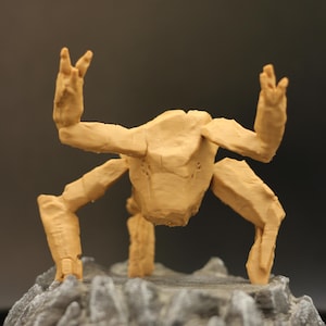 May include: A tan-colored, multi-legged creature figurine with a textured, rock-like appearance. The figure has two raised, claw-like appendages and stands on a base that resembles a jagged, grey, rocky terrain with a dark, uneven surface.