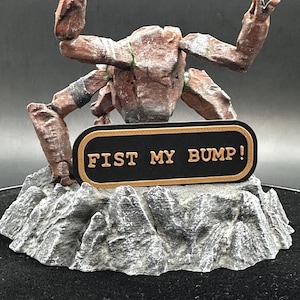 May include: A brown, textured, crab-like creature figurine with raised claws, standing on a gray, rocky base. A black, oval-shaped plaque with a gold border is attached to the front, featuring the embossed gold text FIST MY BUMP!