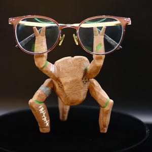 Peut inclure: Un porte-lunettes en bois sculpté en forme de crabe brun, avec deux pinces avant levées soutenant une paire de lunettes à monture brune. La figurine en bois présente des bandes vertes peintes, des accents argentés et des marques blanches.