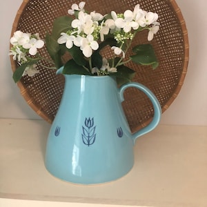 Può includere: Un'anfora in ceramica blu chiaro con un motivo floreale blu e un arrangiamento di fiori bianchi all'interno. L'anfora ha un manico e un'ampia apertura. L'anfora è appoggiata su uno scaffale davanti a un cesto intrecciato.