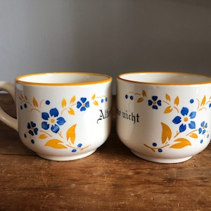 May include: Two white ceramic mugs with handles, featuring blue floral designs and the text "Alte ne nicht". The rims and floral accents are yellow. The mugs are on a wooden surface.