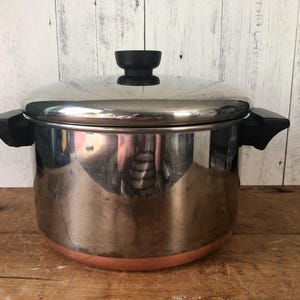 Op de afbeelding: Een roestvrijstalen soeppan met een gepolijste afwerking en een koperkleurige bodem. De pan is voorzien van twee zwarte zijhandgrepen en een bijpassend roestvrijstalen deksel met een zwarte knop aan de bovenkant, die de omgeving reflecteert.