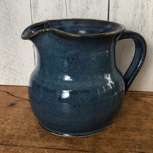 May include: A blue ceramic pitcher with a rounded body and a curved handle. The pitcher has a spout for pouring and a dark rim. The glaze has a speckled appearance, and the pitcher sits on a wooden surface.