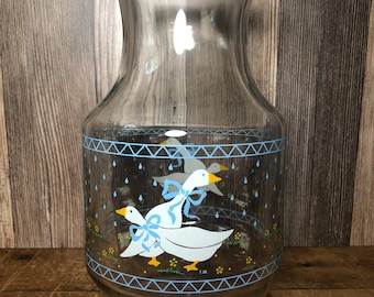 May include: A clear glass vase featuring a decorative wrap-around print of white geese with yellow beaks and blue bows. The design includes blue geometric borders, small yellow flowers, and blue raindrops, with the letters T.M. printed near the base.