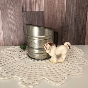 Pode incluir: Uma peneira de farinha de metal vintage com uma alça preta e um botão verde, ao lado de uma pequena estatueta de cavalo de cerâmica. A peneira é prateada e tem uma aparência desgastada. O cavalo é branco com detalhes marrons, sobre uma toalha de renda branca.