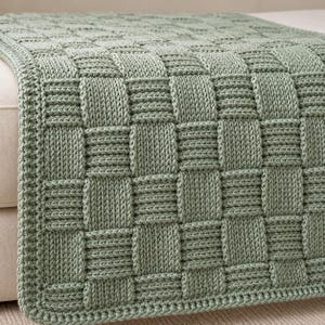 May include: A sage green knitted throw blanket featuring a basketweave pattern. The textile displays a repeating square grid design created by alternating vertical and horizontal knit stitches, resulting in a textured, uniform surface.