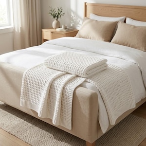 May include: A cream-coloured waffle-knit throw blanket folded neatly on top of a white duvet cover. The blanket features a textured grid pattern and is draped over a bed with a light beige upholstered frame and two matching beige pillows.