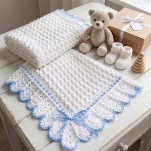 Può includere: Una coperta per neonati all'uncinetto bianca con un motivo a cialda testurizzato, caratterizzata da un bordo smerlato azzurro e un fiocco in nastro azzurro abbinato. Sono inclusi un orsacchiotto di peluche beige, scarpine in maglia bianca e un gioco.