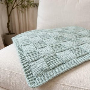 May include: A light sage green knitted blanket featuring a textured basketweave pattern. The square textile has a distinct border with a ribbed edge detail, showcasing a consistent knit stitch throughout the entire surface of the fabric.