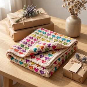Può includere: Una coperta all'uncinetto piegata caratterizzata da un motivo ripetuto di piccoli cuori multicolori nelle tonalità del rosa, viola, arancione, giallo, verde e blu, su uno sfondo di filato color crema con un bordo coordinato.