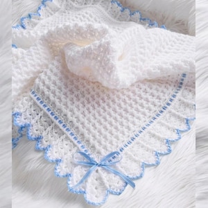 May include: A white crocheted blanket featuring a textured waffle stitch pattern. The edges are finished with a scalloped border in light blue, and a light blue ribbon is woven through the border, tied into a small bow at the corner.