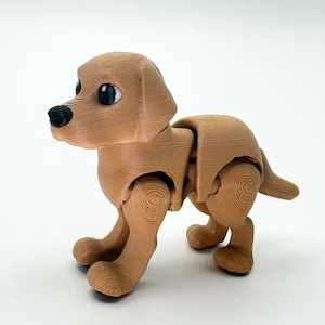 May include: A light brown, articulated dog figurine with a textured, wood-like appearance. The dog has black eyes and nose, and its legs and body are jointed. The tail is small and upright. The figurine is standing on a white surface.