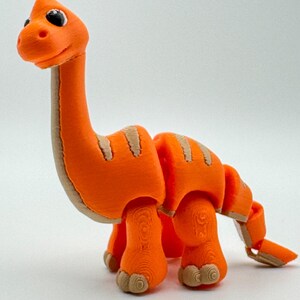 Może przedstawiać: Pomarańczowa, przegubowa zabawka dinozaura z długą szyją i ogonem. Zabawka ma beżowe paski i akcenty, czarne oczy i beżowe stopy. Dinozaur stoi na białej powierzchni.