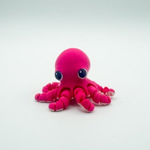 May include: A small, bright pink octopus figurine with large, dark blue eyes. The octopus has eight curled tentacles with white tips. The figurine is set against a plain white background.