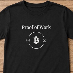 Könnte beinhalten: Ein schwarzes Kurzarm-T-Shirt mit dem weißen Schriftzug Proof of Work, der über einer kreisförmigen Grafik zentriert ist. Die Grafik zeigt ein Bitcoin-Symbol, das von zwei kleinen Würfeln flankiert wird, alles in klarem weißem Liniendesign.