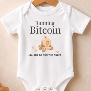 Könnte beinhalten: Ein weißer kurzärmeliger Babybody mit dem Text Running Bitcoin und Raised to Run the Rules. Die Grafik zeigt ein hölzernes Spielzeugauto mit einem Bitcoin-Symbol an der Seite und kleinen Fußabdrücken, gedruckt in Dunkelgrau und Hellbraun.