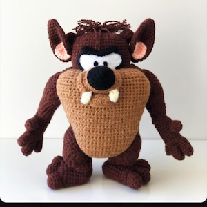 Puede incluir: Un peluche hecho a mano de ganchillo del Demonio de Tasmania. El peluche presenta un cuerpo marrón, vientre beige, nariz negra, ojos blancos y dos dientes blancos. Las orejas tienen detalles en rosa.