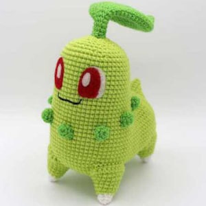 May include: A handmade crochet plush toy in the shape of a green creature with red eyes and a green leaf-like structure on its head. The toy has small green pom-poms and white accents on its feet.