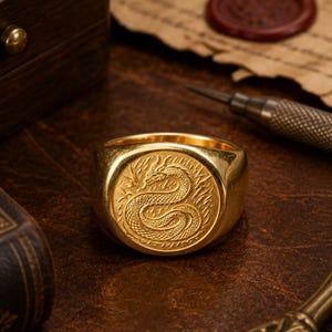 May include: A gold-toned signet ring featuring a circular face engraved with a detailed serpent or dragon motif. The ring has a polished, smooth band and a flat, circular top surface that displays the intricate, textured scales of the creature.