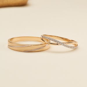 May include: Two gold rings, one wider band featuring a smooth polished finish with a subtle diagonal silver-toned inlay, and one thinner band with a delicate twisted design encrusted with small clear stones along the upper half of the band.