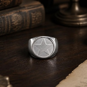 Op de afbeelding: Een zilverkleurige zegelring met een cirkelvormig vlak waarin een gestructureerde vijfpuntige ster in het midden is gegraveerd. De ring heeft een gepolijste, gladde band en een metalen afwerking met een strak ontwerp en een duidelijk stermotief.
