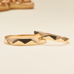 May include: Two gold rings with a faceted, geometric surface design. The larger ring features a small section embedded with tiny clear stones, while the smaller ring has a smooth, polished gold finish with matching angular facets.