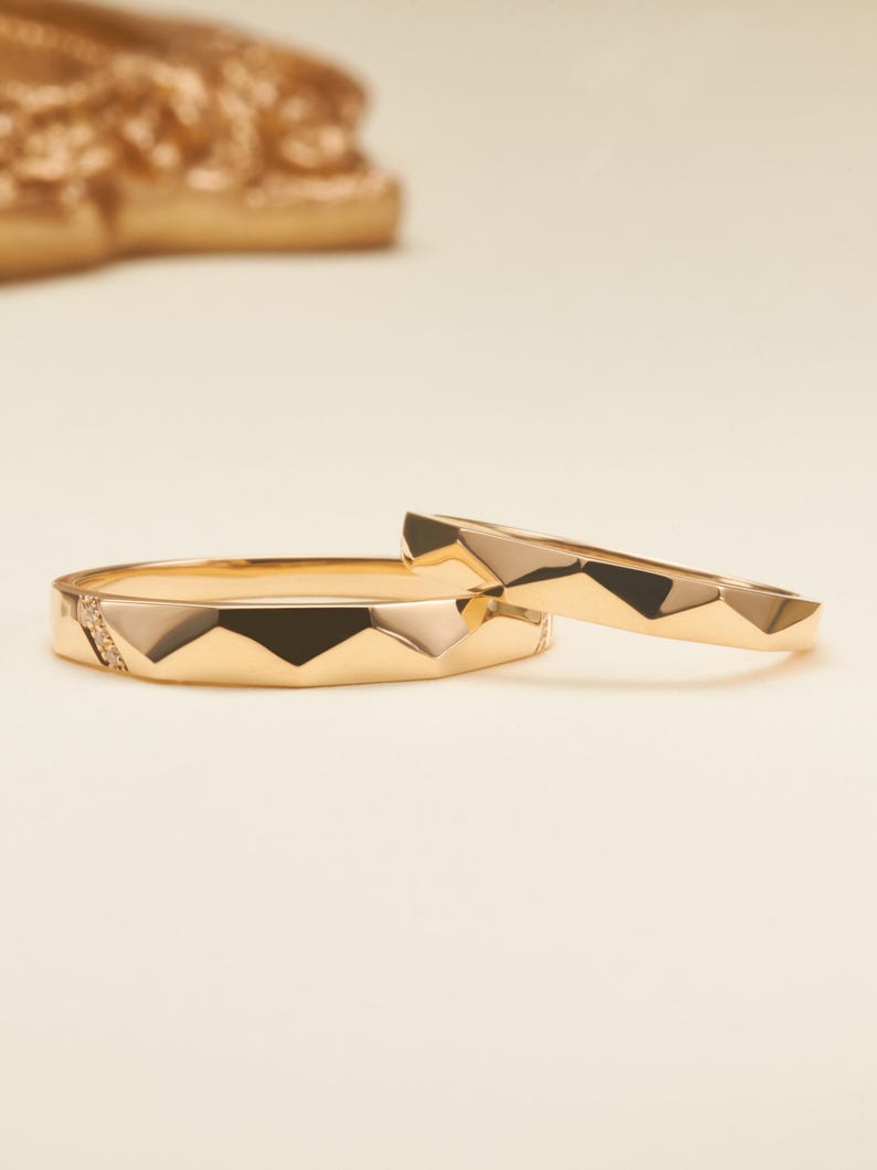 May include: Two gold rings with a faceted, geometric surface design. The larger ring features a small section embedded with tiny clear stones, while the smaller ring has a smooth, polished gold finish with matching angular facets.
